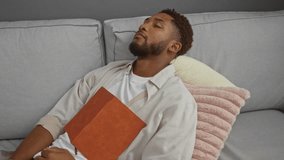 Young man sleeping with a closed book resting on his chest in a cozy living room setting with pillows and a couch - Powered by Shutterstock - Get 15% off with code: PIKWIZARD15