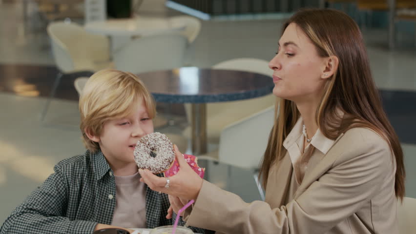 Tilt down shot of happy mother and her teenage son eating donuts while sitting at table with drinks on food court