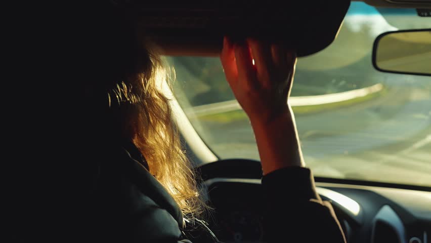 A woman with wavy hair is driving a car. The sun shines in her eyes and she lowers her sun visor. Rear view from the side. Traveling by car during sunrise