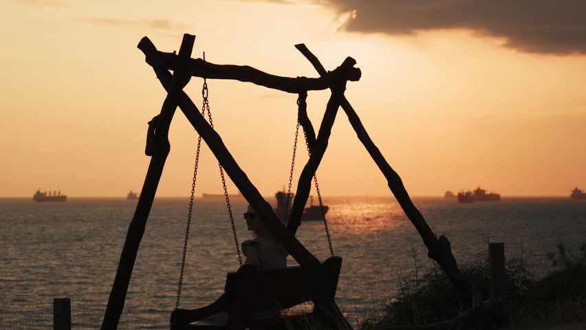 unrecognizable girl swinging on a swing against the backdrop of a sunset and ocean or sea. Silhouette of a young woman taking a break from routine at the edge of the world.