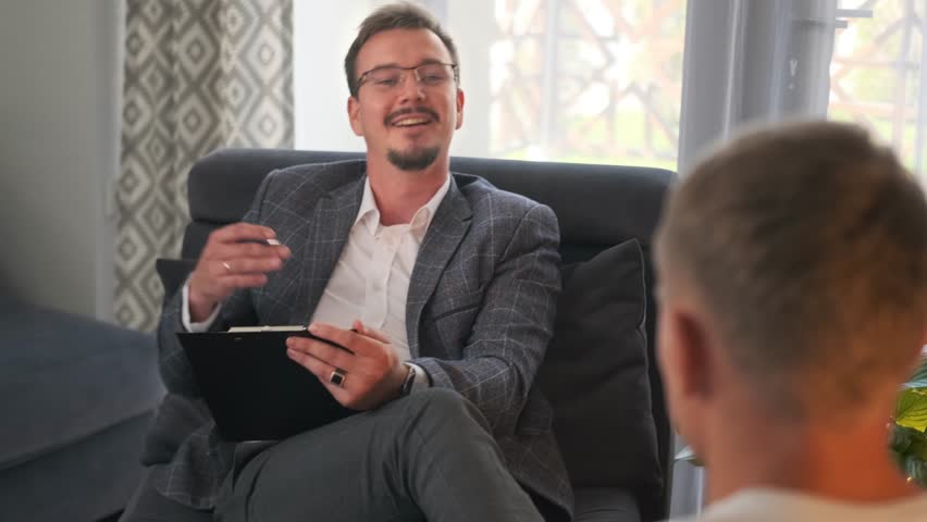 Seated with a clipboard the psychologist smiles warmly, using gestures to explain cognitive behavioral therapy for depression. The patient listens closely, feeling supported and understood. 