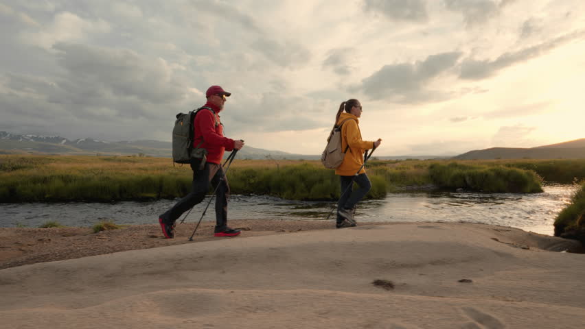 Two adult hikers with backpacks and hiking poles walks at river shore against snowy mountains at sunset time