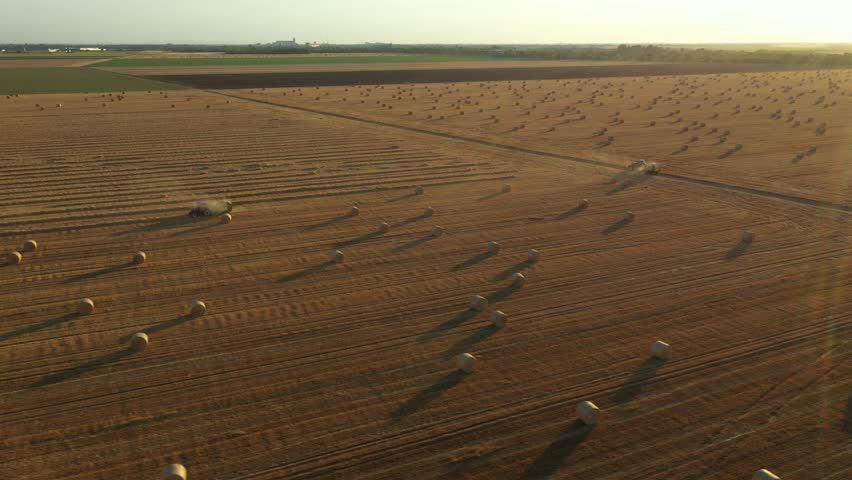 Above view, dolly move orbit with backlight around two tractors as they pulling round balers, machines roll up the straw and spits out a packed round bale over agricultural field, working together