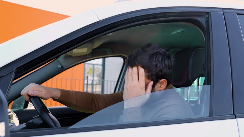 A young man driver stopped while sitting behind the wheel and holds his head with his hand. Better to stop to avoid an accident. The driver is a guy in the car.