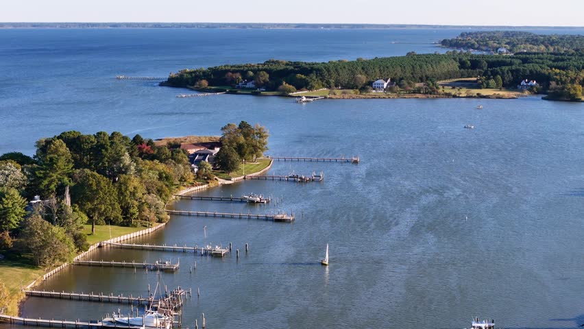 A drone soars over the Chesapeake Bay, capturing a sailboat gliding across the blue water, surrounded by lush green trees, creating a peaceful and scenic coastal view.