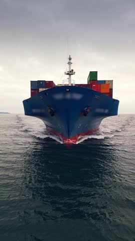 Front view of a blue container ship sailing through calm waters under a cloudy sky, heading towards the port