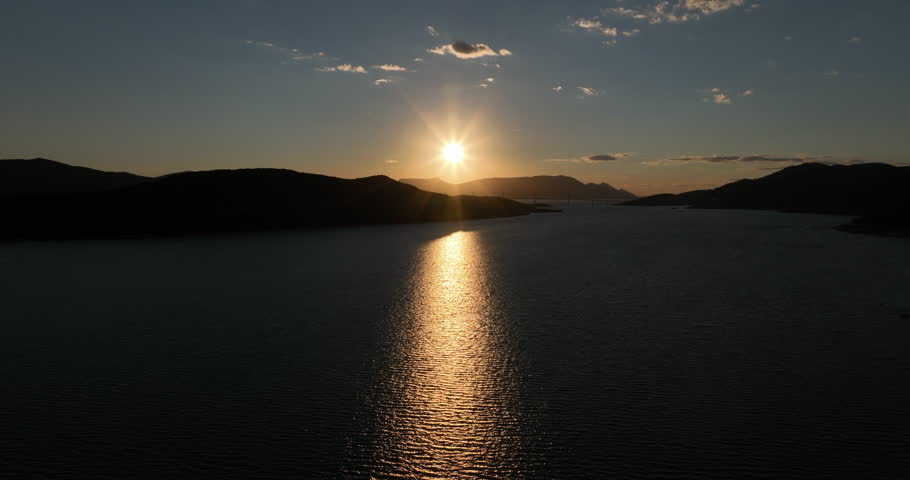 A breathtaking sunset scene captured from a high vantage point. The sun dips below the horizon, casting a warm golden glow on the tranquil water. Dark silhouettes of mountains frame the scene.