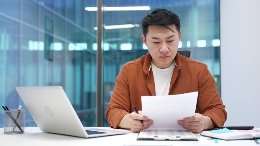 Confused asian businessman having difficulty with paperwork sitting at a desk at workplace in business office. Puzzled male worker checking documents on computer and cannot understand problem