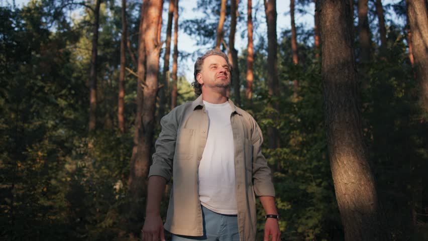 Camera focus on Caucasian man in white t-shirt and grey shirt standing in middle of forest. Lookin up and making open arms gesture. Warm sunshine shining on persons face. Green trees around.