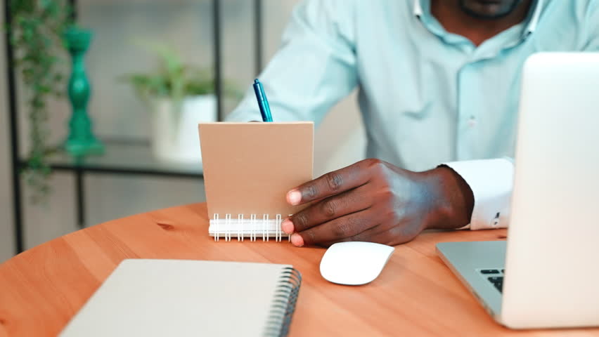 Attentive serious african american freelancer man makes notes writes important information in notepad looking at laptop screen sits in home office working online. Digital nomad, remote job concept.