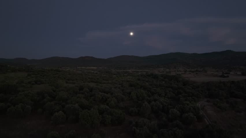 ascending flight with a drone at dusk, a group of trees appears, the field and we see the line of mountains in the background, in the sky the moon appears behind some clouds, it is fantastic
