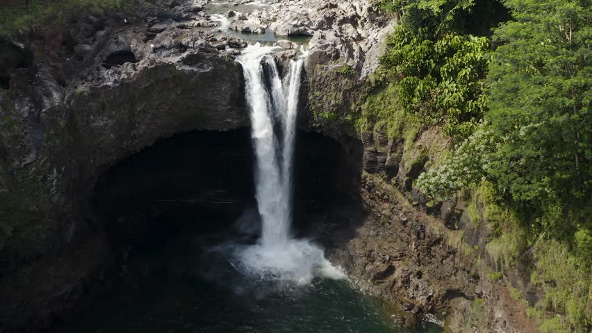 A serene waterfall plunging into a natural pool, surrounded by lush greenery and rocky cliffs under bright daylight.
