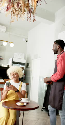 Vertical video: Barista in apron taking payment from woman with smartphone in cafe. Coffee, transaction, technology, slow motion