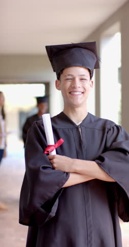 Vertical video: Teenager in graduation gown holding diploma, smiling proudly in school hallway. achievement, education, celebration, success, slow motion