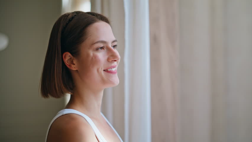 Smiling woman pulling curtains enjoying serene morning routine closeup. Reflective lady letting sunlight in modern living room creating cozy atmosphere. Happy brunette looking window starting new day.