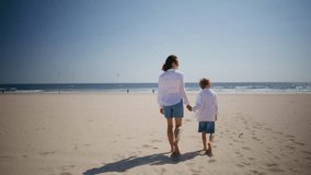 Happy woman walking with son on beautiful beach under summer sun. Relaxed mother child strolling barefoot at sandy seashore holding hands together. Carefree family enjoying sunny vacation on seacoast. - Powered by Shutterstock - Get 15% off with code: PIKWIZARD15