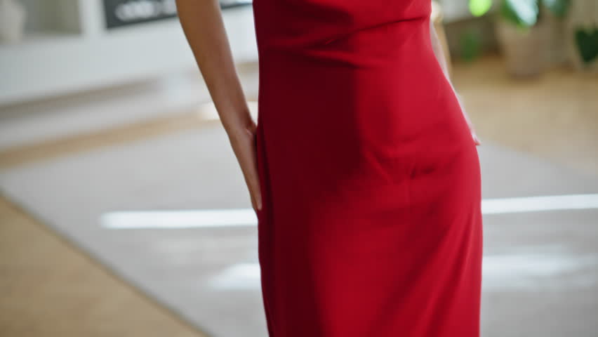 Glamorous woman in red dress posing in large mirror admiring elegant outfit closeup. Stylish lady moving gracefully enjoying reflection. Chic brunette showcasing fashion clothing at home apartment.
