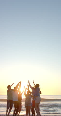 Vertical video: Group of friends enjoying sunset, raising hands and celebrating on beach. Friendship, celebration, outdoors, vacation, togetherness, slow motion