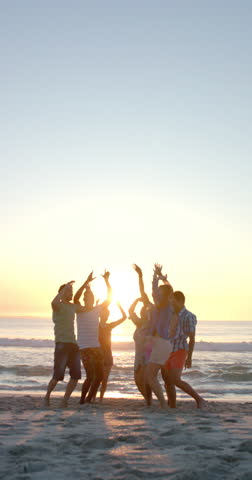 Vertical video: Group of friends enjoying sunset, raising hands and celebrating on beach. Friendship, celebration, outdoors, vacation, togetherness, slow motion