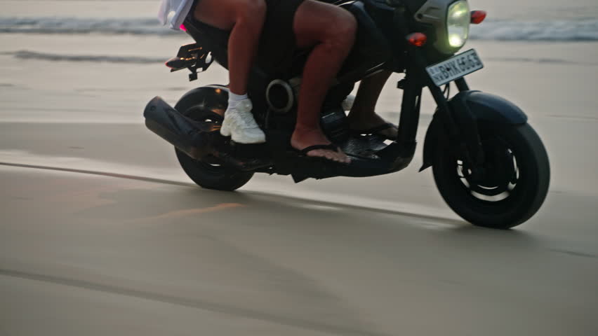 Travel couple ride motorcycle along sea beach on sunrise. Intraframe installation from bike wheels reflection, trace to people. Tourists drive rented motorbike on ocean coastline. Woman hugs man.