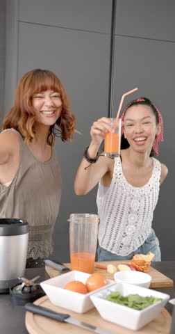 Vertical video: Smiling women in kitchen making fresh juice with blender and enjoying drinks. Cooking, juicing, healthy lifestyle, domestic life, nutrition, happiness, slow motion