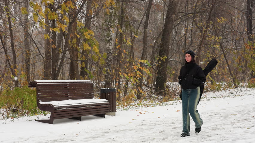 Music artist in winter jacket with hands in pockets and guitar on back walking in snowfall, snowy bench and dry trees in background, serene winter landscape