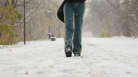 Low angle view of someone in black boots and jeans, carrying an item behind them while walking on a snowy path during snowfall, with trees and a bench in the serene winter landscape - Powered by Shutterstock - Get 15% off with code: PIKWIZARD15