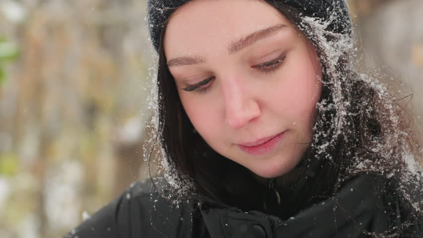 Close-up of a young woman in black beanie with snowflakes on her hair and jacket, glancing upward with a warm smile before looking away, capturing a contemplative moment amidst soft falling snow