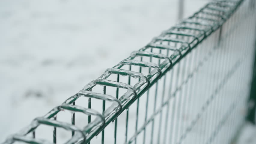 Close-up of green metal fence coated with ice, showcasing icicles hanging from the wires against a blurred snowy park background, emphasizing the stark contrast between frozen textures