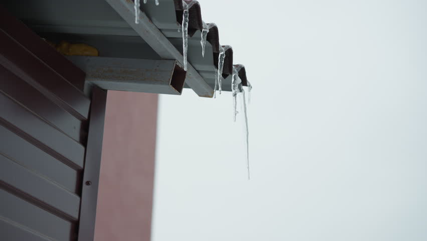 Close-up of roof edge featuring frosted metal structure with icicles hanging down, highlighting frozen details and icy textures, set against a minimalistic winter sky with a blurred urban background