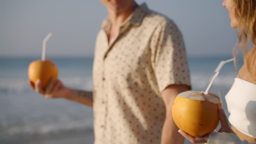 Romantic couple drink coconut water walking on sea beach. People sip alcohol cocktails from straws on vacation. Man, woman relax, quench thirst with healthy superfood beverage. Slow motion, close-up.