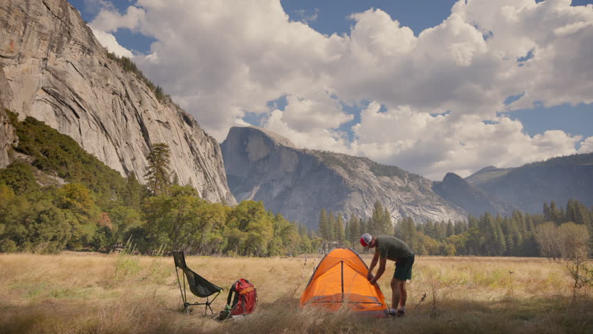A hiker is setting up a vibrant orange tent in the breathtaking landscape of Yosemite National Park, which features majestic and iconic rock formations along with a peaceful atmosphere to enjoy