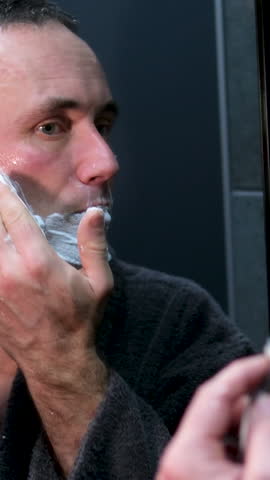 man in robe after shower spa treatments near mirror shaving foam beauty salon self-care at home bathroom man in front of a mirror shaves his face Serious older man looking in his reflection in mirror
