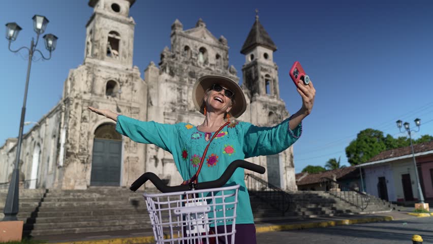 A smiling woman takes a selfie with her phone and bike in Granada, Nicaragua, near the historic Iglesia Guadalupe church. She expresses her happiness while embracing the outdoor experience.
