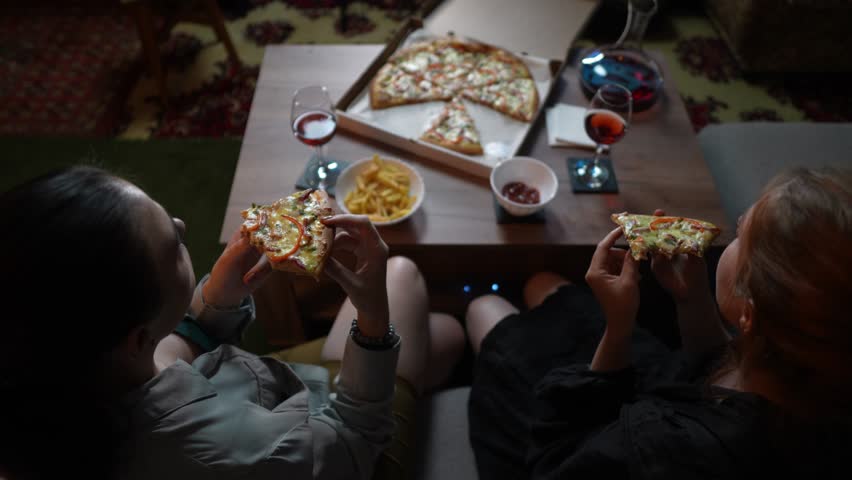 Back view of two women eating pizza and watching TV in the evening at home. Slow motion of friends spending weekend in front of film with junk food. Theme of leisure time activity and tasty dinner