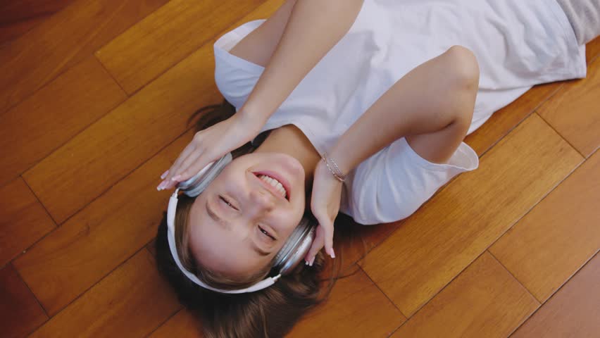 A relaxed woman is enjoying her music while wearing comfortable headphones, lounging on a warm wooden floor, embodying a carefree and chill atmosphere perfect for unwinding after a long day