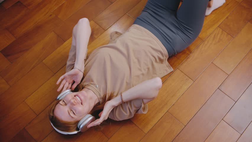 A relaxed woman enjoys calming music on a wooden floor, embracing the cozy vibes of her artistic space. Immersed in leisure, she feels content and happy, smiling in blissful enjoyment