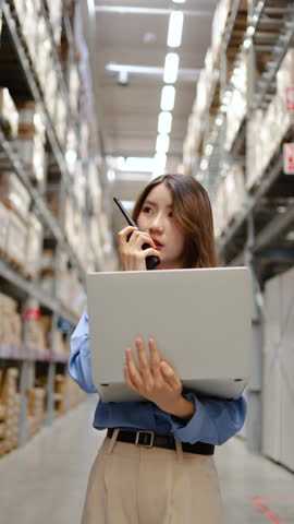 lively warehouse, dedicated young asian woman meticulously checks inventory details on her laptop while engaged in a conversation for radio communication, ensuring smooth operations and accuracy