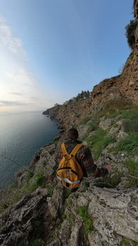 Vertical video. A Male Traveler With a Yellow Backpack Carefully Walking Down a Rocky Cliffside Path Toward the Ocean, Surrounded by Rugged Terrain and Vegetation. Coastal Trekking