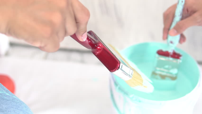 Hand holding a paintbrush and dipping it in the bucket of blue paint in order to paint the wall in home.