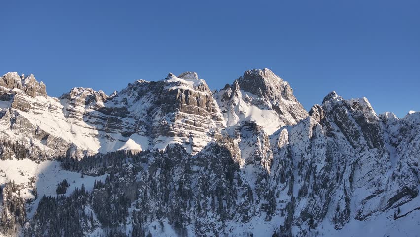 oaring aerial view showcases the majestic Fronalpstock mountain range in Glarus, Switzerland. Snow-capped peaks pierce a brilliant blue sky, creating a breathtaking winter landscape.