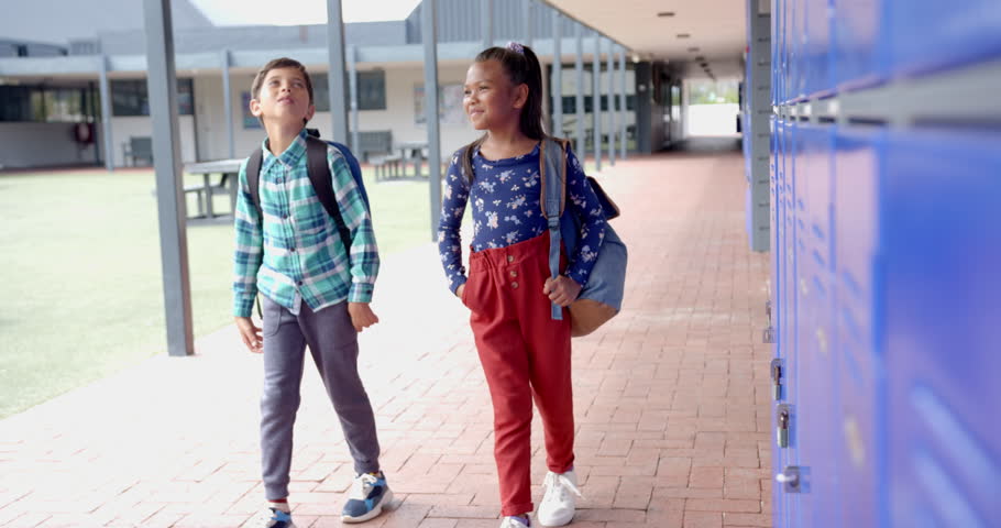 In school, two biracial students are walking by blue lockers. Boy wearing plaid shirt and girl in star-patterned top laughing together, both with backpacks, unaltered, slow motion