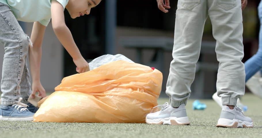 In school, child picking up trash with large orange bag, cleaning playground. Volunteering, environment, community, responsibility, childhood, unaltered, slow motion