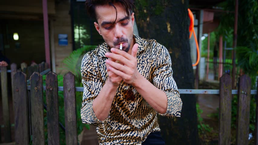 Man stands outside the restaurant lighting cigarette, Guy enjoys smoke while standing outdoors, Young male leans against wooden fence, Person puffing a cigarillo, Holding his fag, Rolled tobacco, Puff