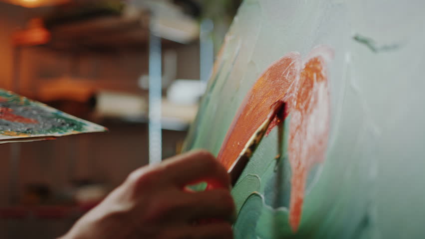 Detailed view of hand of unrecognizable painter using brush to apply rich orange paint to canvas in warmly-lit art studio