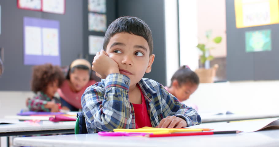 In school, boy daydreaming at desk while classmates and teacher working. Education, classroom, learning, childhood, students, distraction, unaltered, slow motion