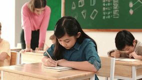 Asian girl writing test or classwork while looking at camera at classroom. Diverse happy student taking notes and teacher help caucasian boy doing test while sitting in front of blackboard. Pedagogy. - Powered by Shutterstock - Get 15% off with code: PIKWIZARD15