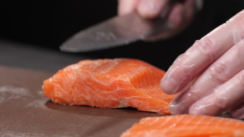 Macro. The chef's hand in a transparent glove cuts the salmon meat into thin pieces with a knife and puts them in a pile in the foreground. Brown cutting board. Making sushi.