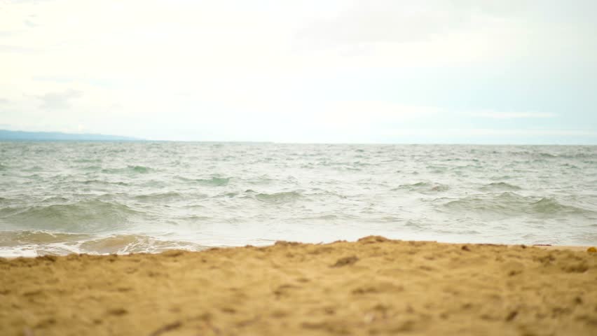 Slow motion view of small waves rolling onto the sandy beach under a cloudy sky in Costa Rica. The scene showcases harmonious colors of gold, blue, and grey.