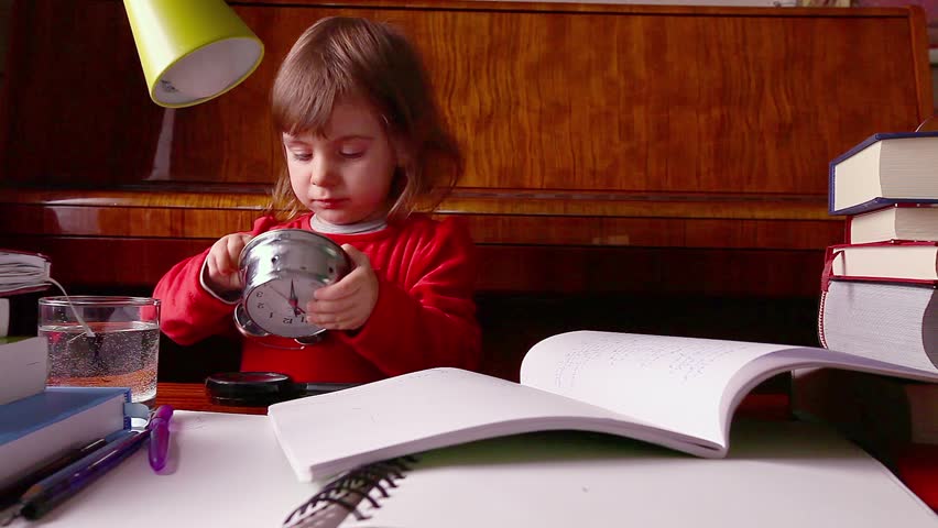 Little baby girl winds up clock and is surrounded by books.
